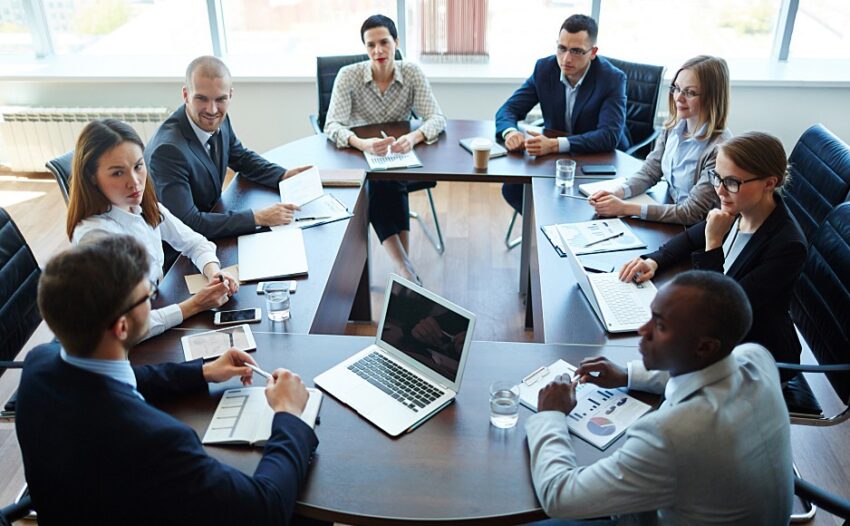 A diverse group of business professionals engaged in a meeting around a conference table with laptops and documents.