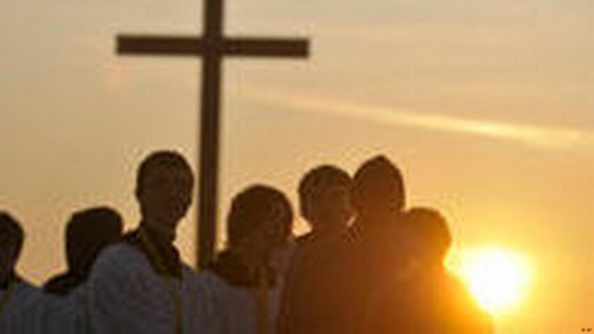 Group of people standing near a large cross at sunset, silhouetted against the glowing sky.