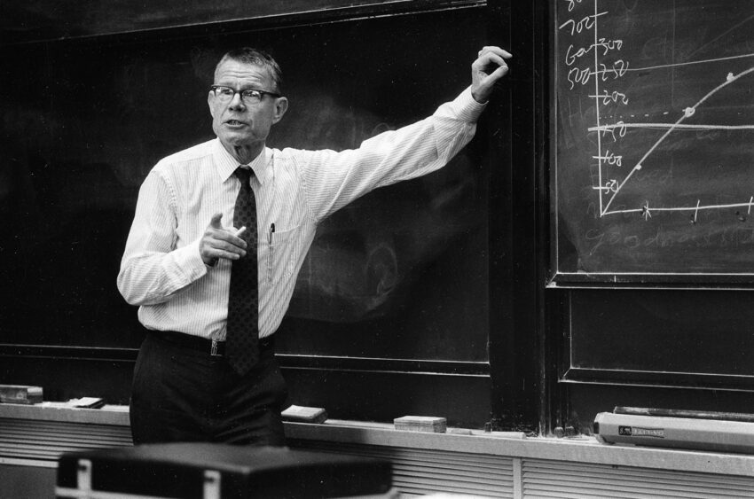 A male professor or teacher stands by a chalkboard, pointing at the board while holding a piece of chalk in a classroom.