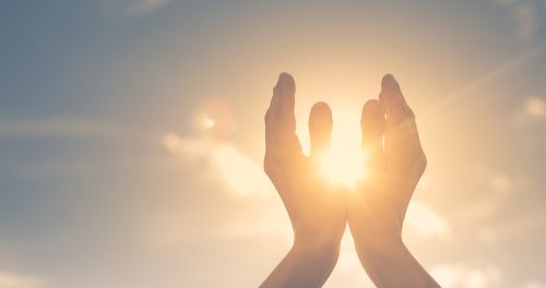 Hands gently cupping the bright sun against a soft, glowing sky during sunset or sunrise.