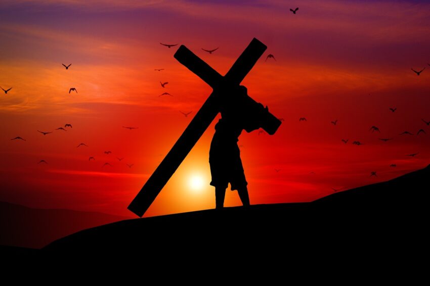 Silhouette of a person carrying a large cross against a vibrant red and orange sunset sky.