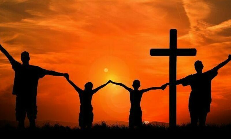 Silhouettes of two people holding hands near a cross against a vibrant orange sunset.