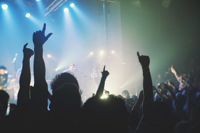 Crowd at a concert with hands raised, illuminated by blue stage lights and smoke effects.