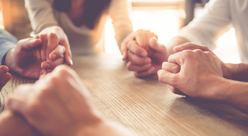People holding hands together around a wooden table in a warm, supportive gesture.